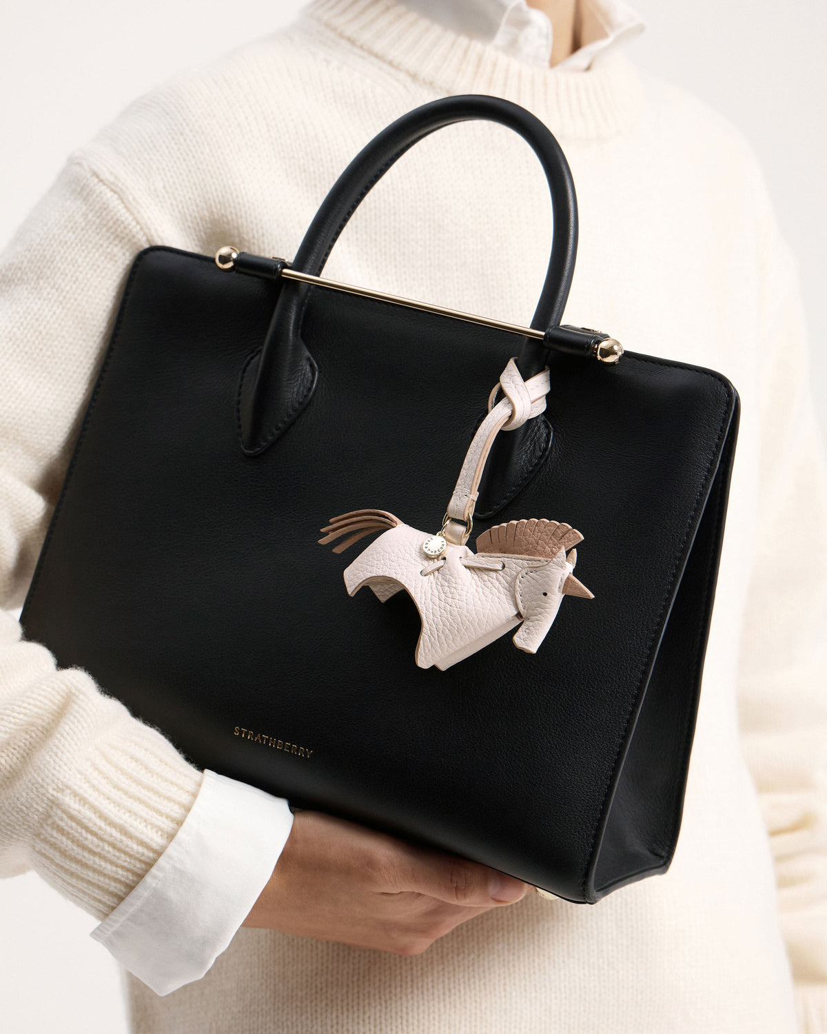 White peony unicorn bag charm on black Strathberry leather bag held by model.